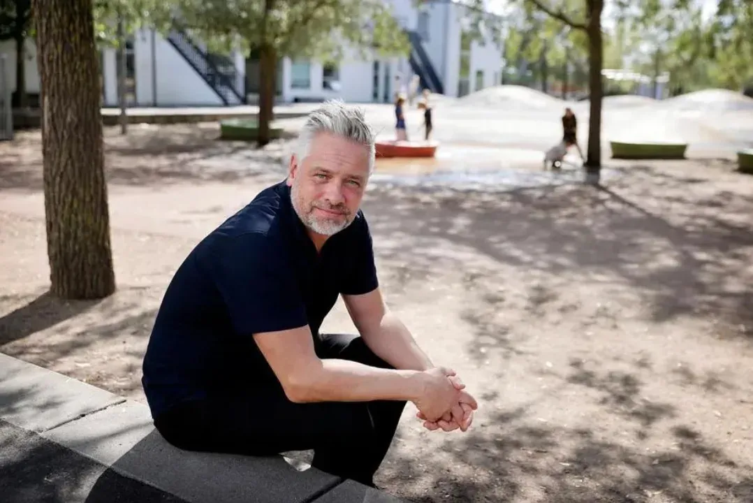 Lars Bechager sidder på en kant i skolegården, mens børn leger i baggrunden.
