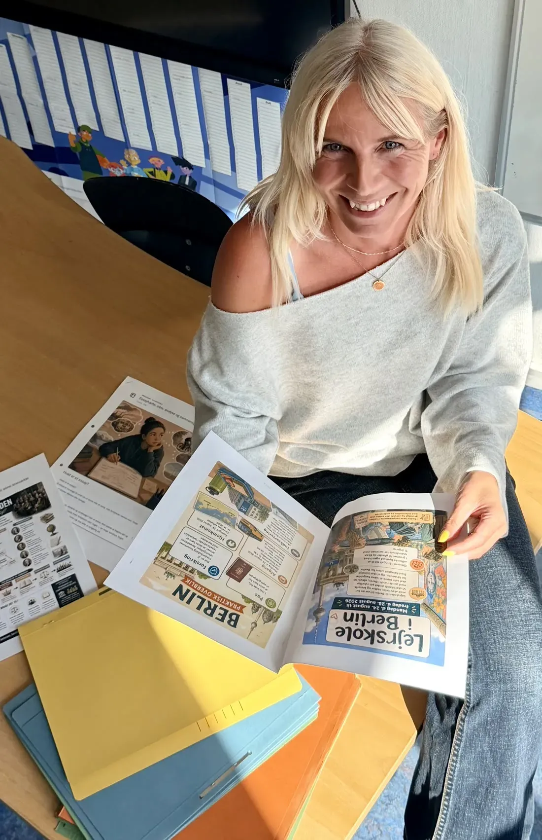Mette Brink smiler op mod kameraet med illustrerede undervisningsmaterialer og farvede mapper på bordet foran sig.